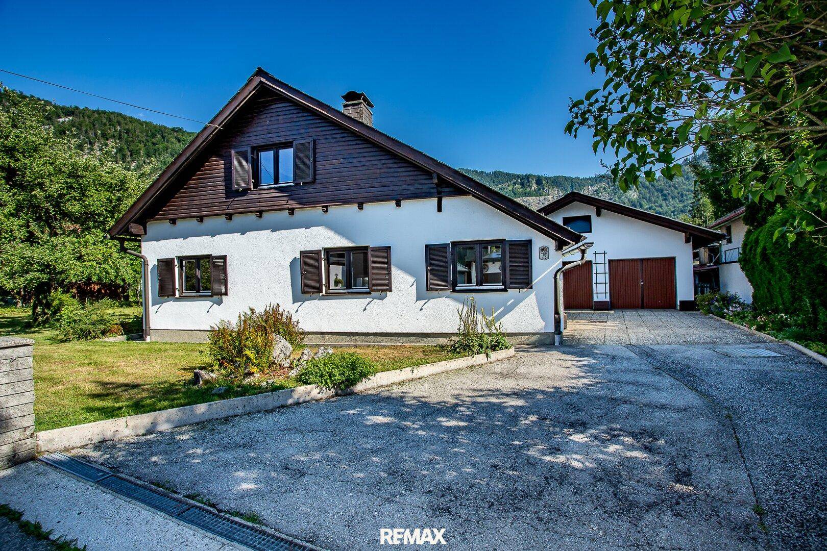 Ansicht des Hauses mit weißer Fassade, braunen Fensterläden und angrenzender Garage mit Zufahrt.