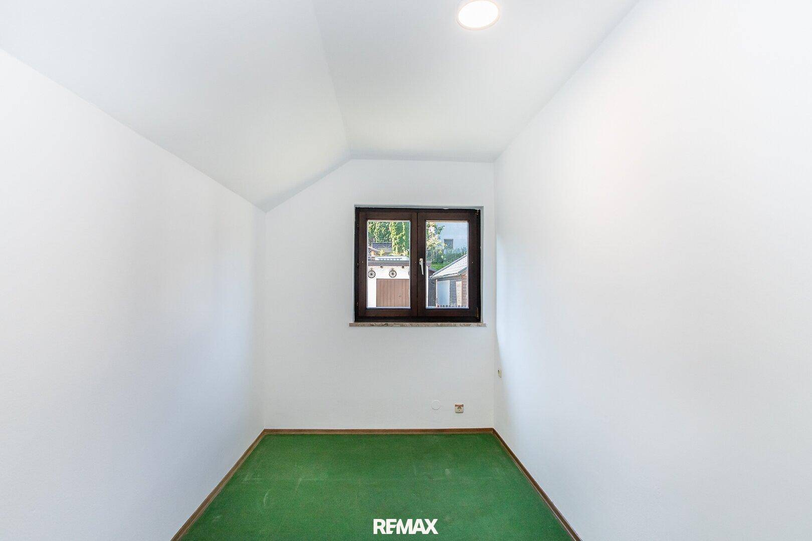 Leeres Zimmer mit grüner Auslegware, weißen Wänden und einem Fenster mit Blick nach draußen.