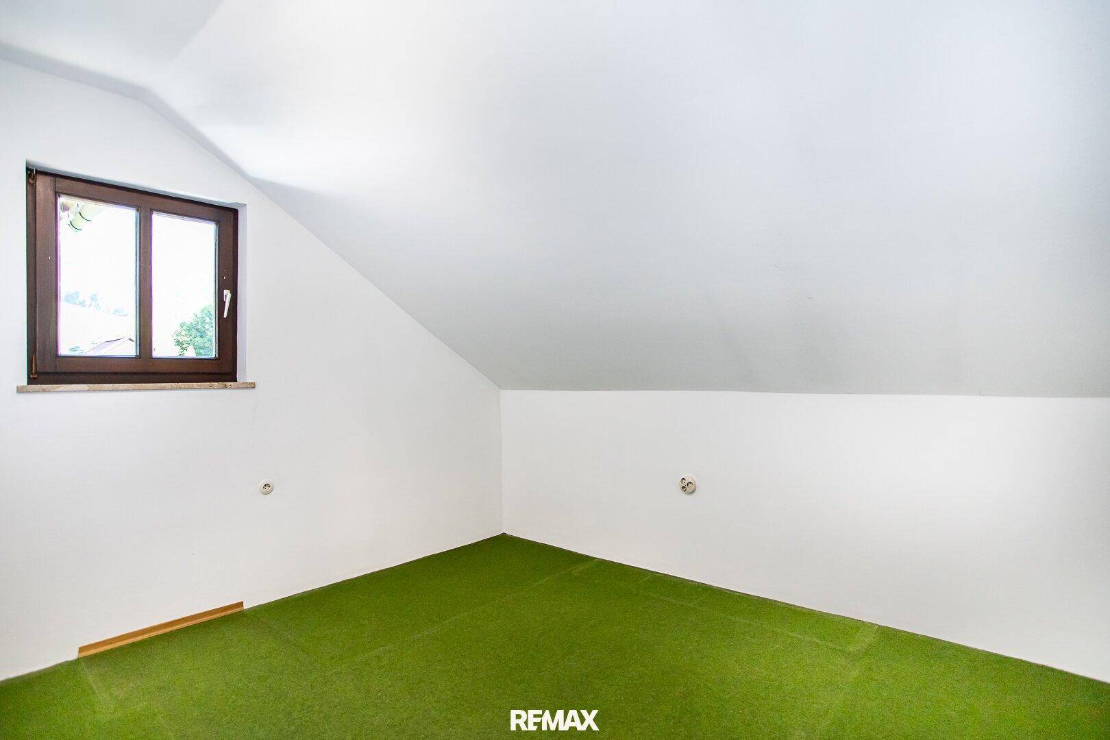Dachzimmer mit schrägen Wänden, grüner Auslegware und einem Fenster, das viel Licht hereinlässt.