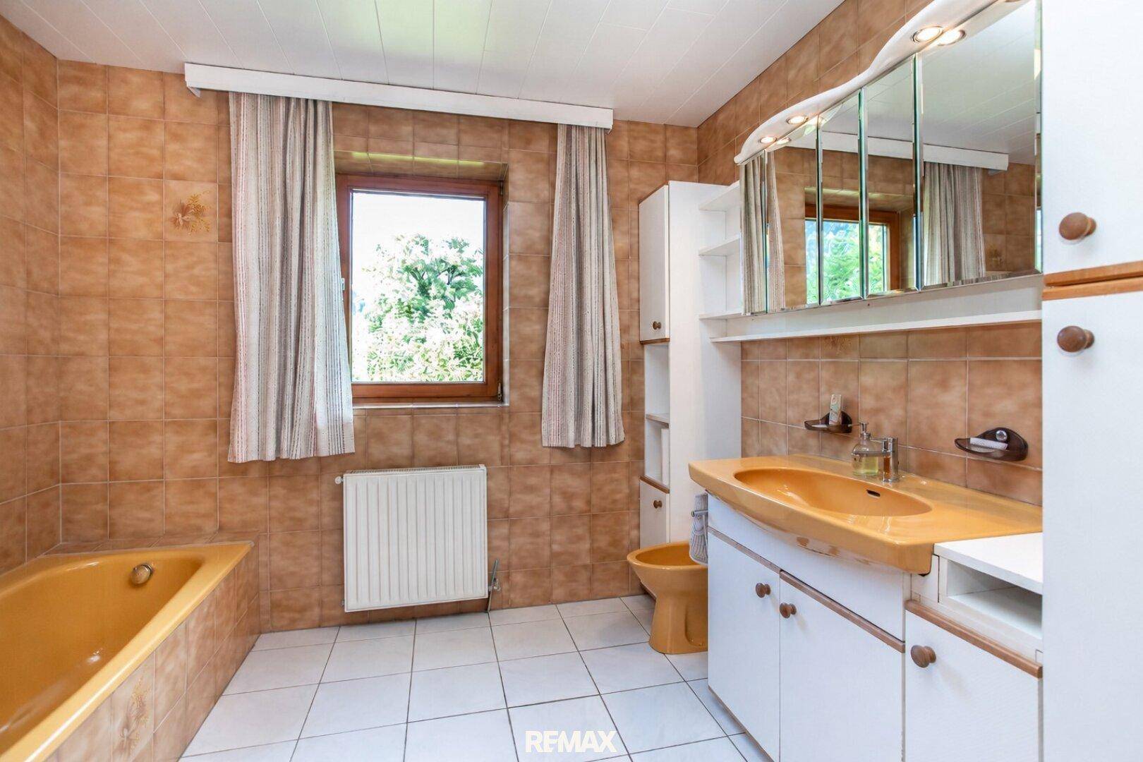 Badezimmer im 80er-Jahre-Stil mit braunen Fliesen, gelber Badewanne und Fenster.