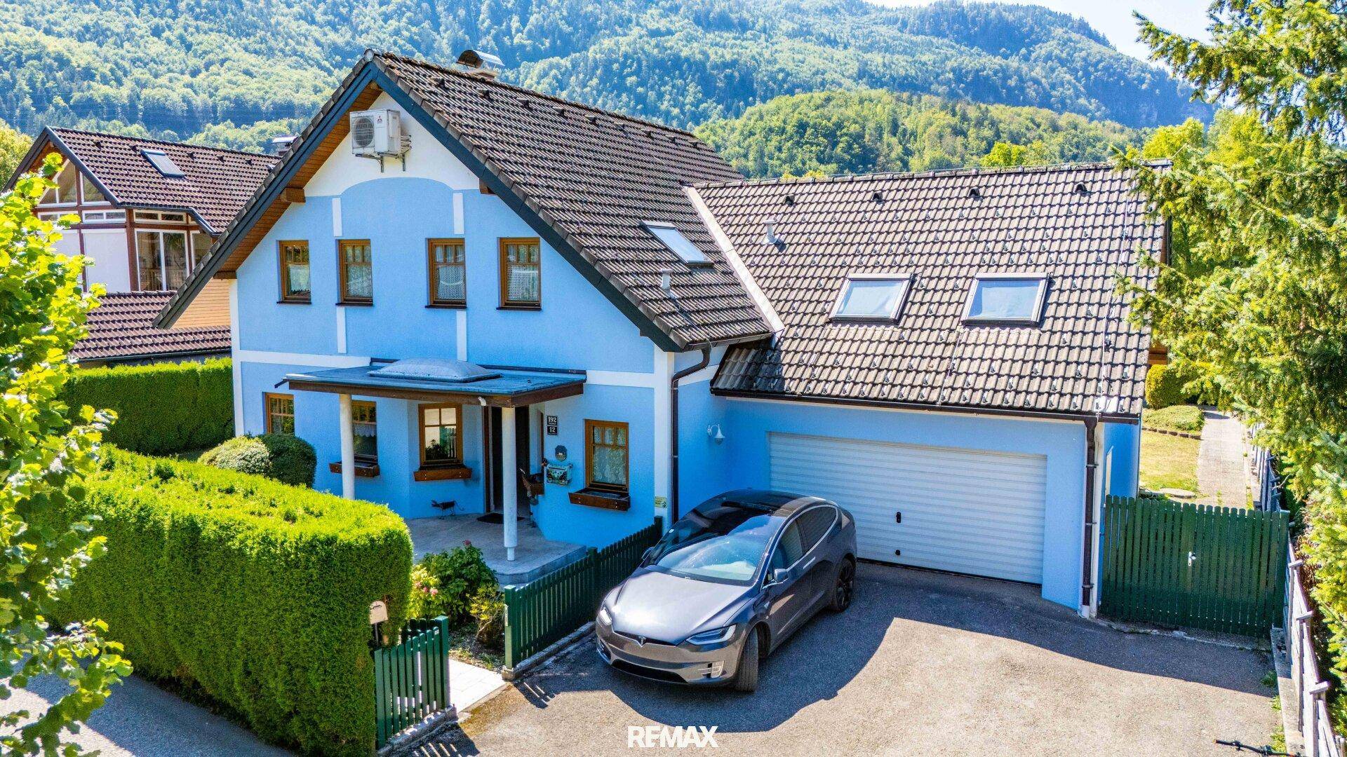 Vorderansicht des blauen Einfamilienhauses mit Garage, Auffahrt und Bergblick im Sonnenschein.