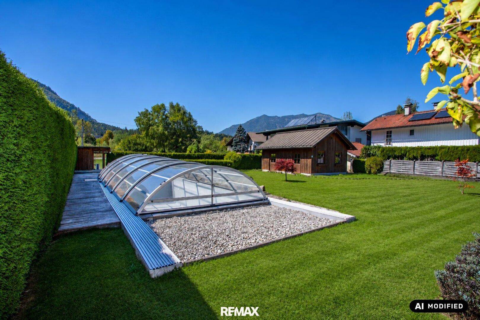 Großer Garten mit Rasenfläche, Hecke und überdachtem Pool vor Bergkulisse.