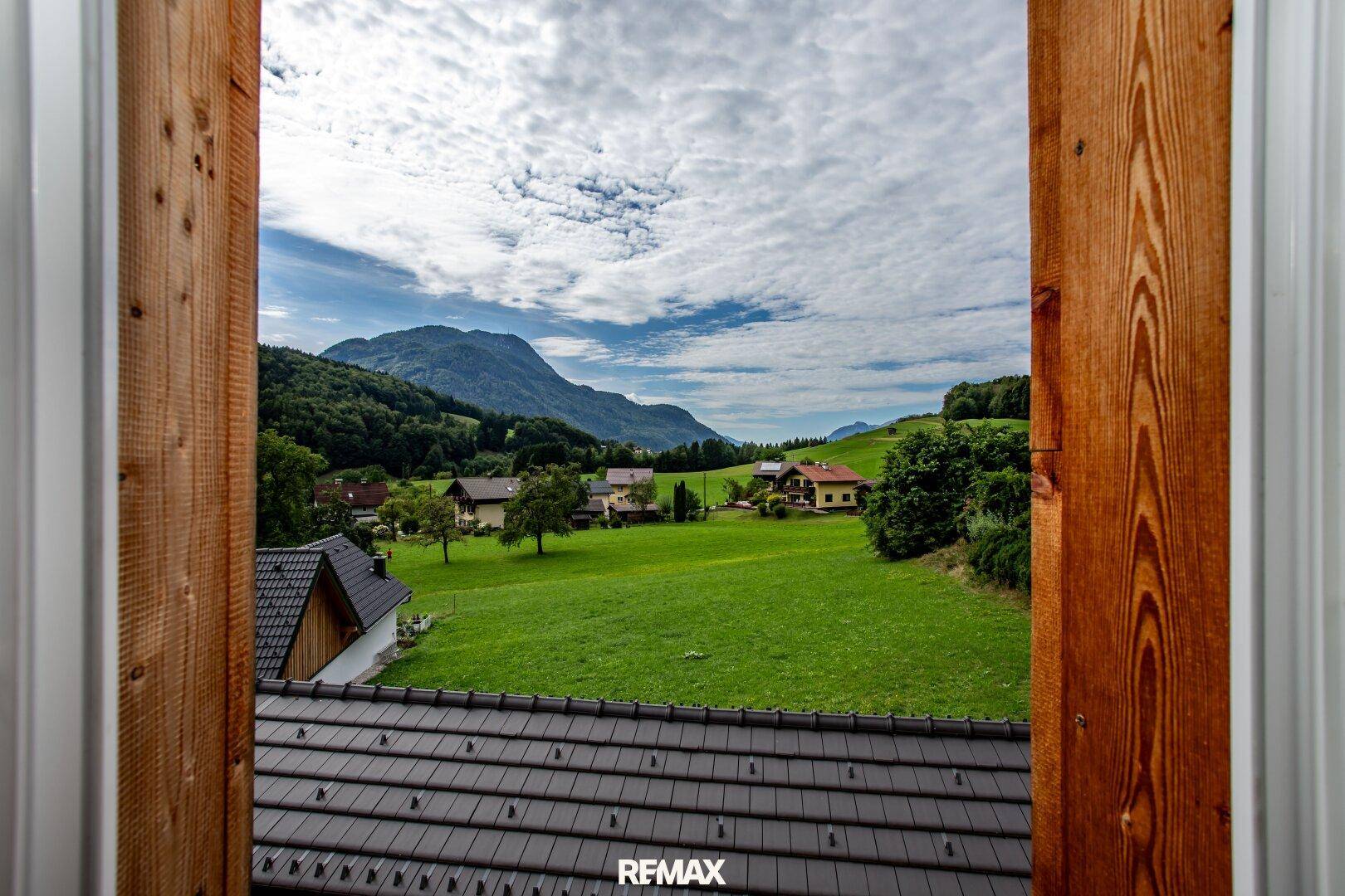 Malerischer Ausblick aus dem Fenster auf grüne Wiesen, Häuser und die umliegenden Berge.
