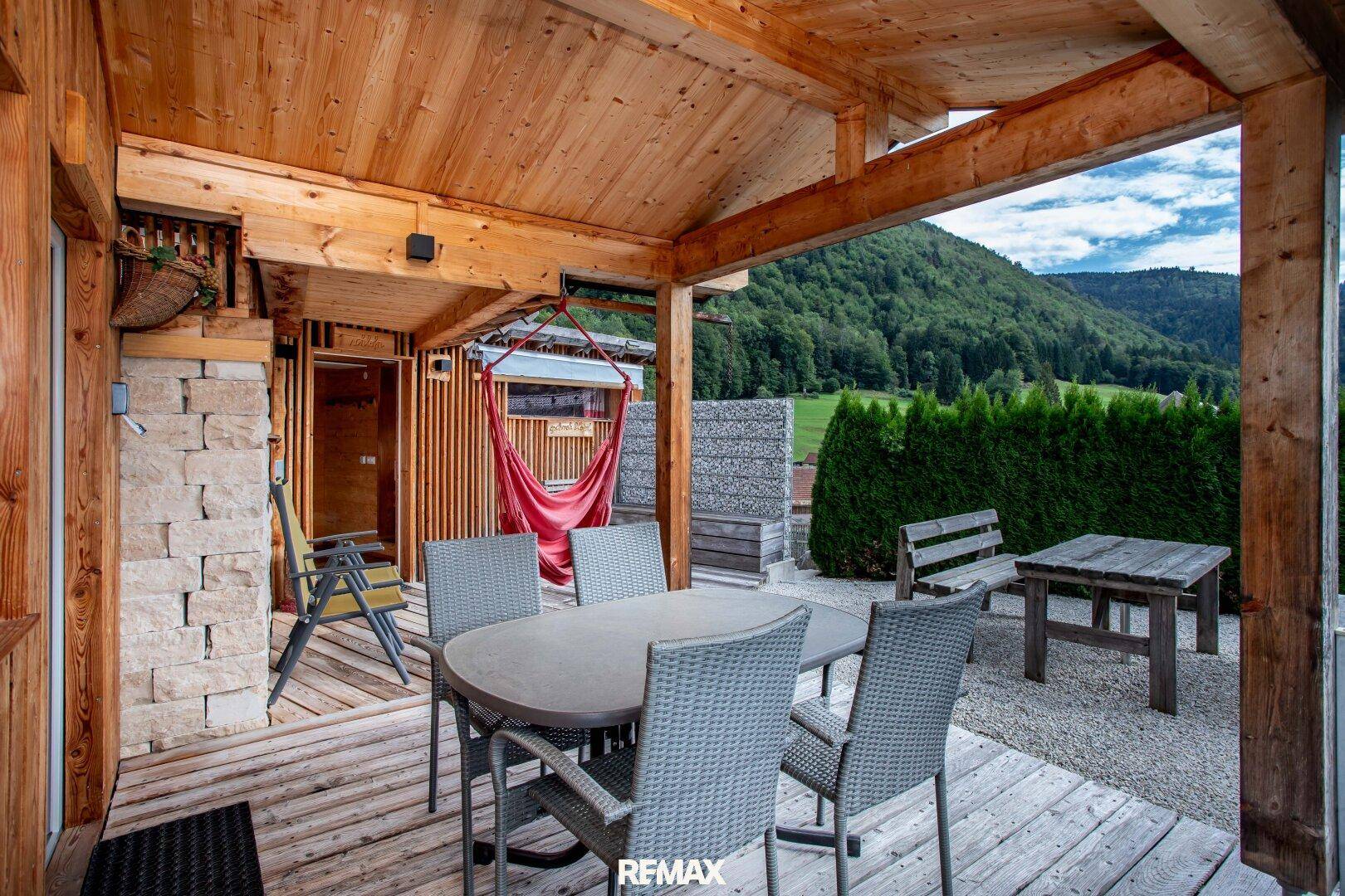 Überdachte Terrasse mit Esstisch und Stühlen, umgeben von grünen Bergen und Natur.