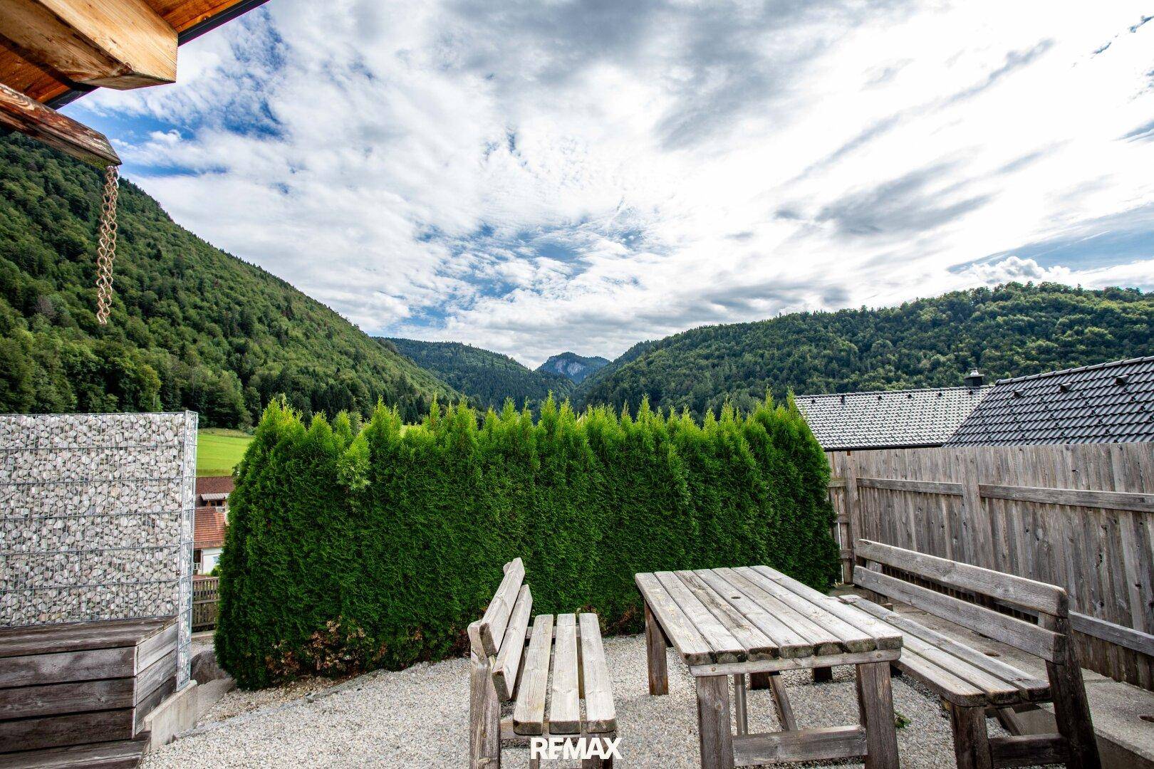 Gartenbereich mit rustikalen Holzmöbeln, Hecke und weitem Blick auf die Berge und Landschaft.