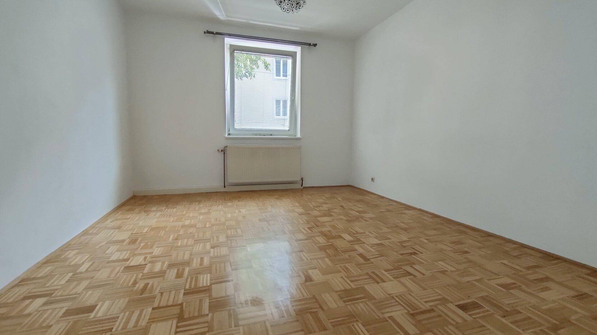 Geräumiges Schlafzimmer mit hellem Parkettboden und einem Fenster mit Blick auf ein Nachbargebäude.