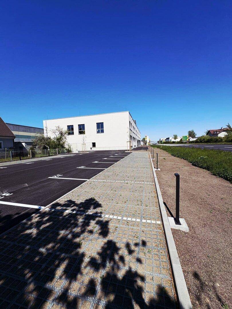 Weitwinkelaufnahme des modernen weißen Gebäudes mit großem Parkplatz und Schattenwurf auf der Gitterpflasterung.