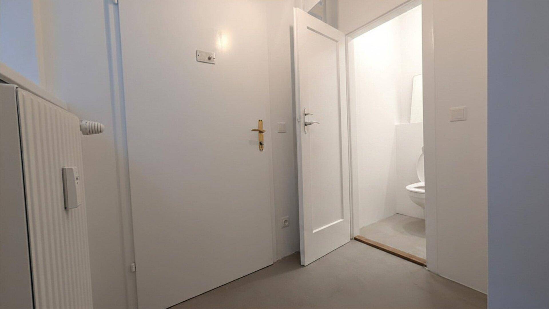 Blick vom Flur in eine Toilette, mit einer weißen Tür, einem Heizkörper und hellem Bodenbelag.