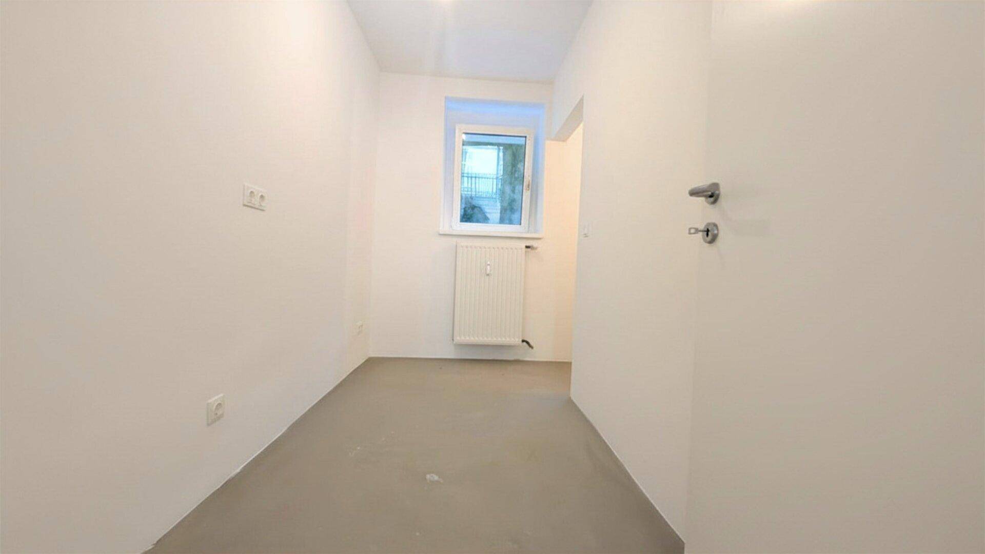 Kleiner, leerer Raum mit weiß gestrichenen Wänden, einem Fenster und einem Heizkörper unter dem Fenster.