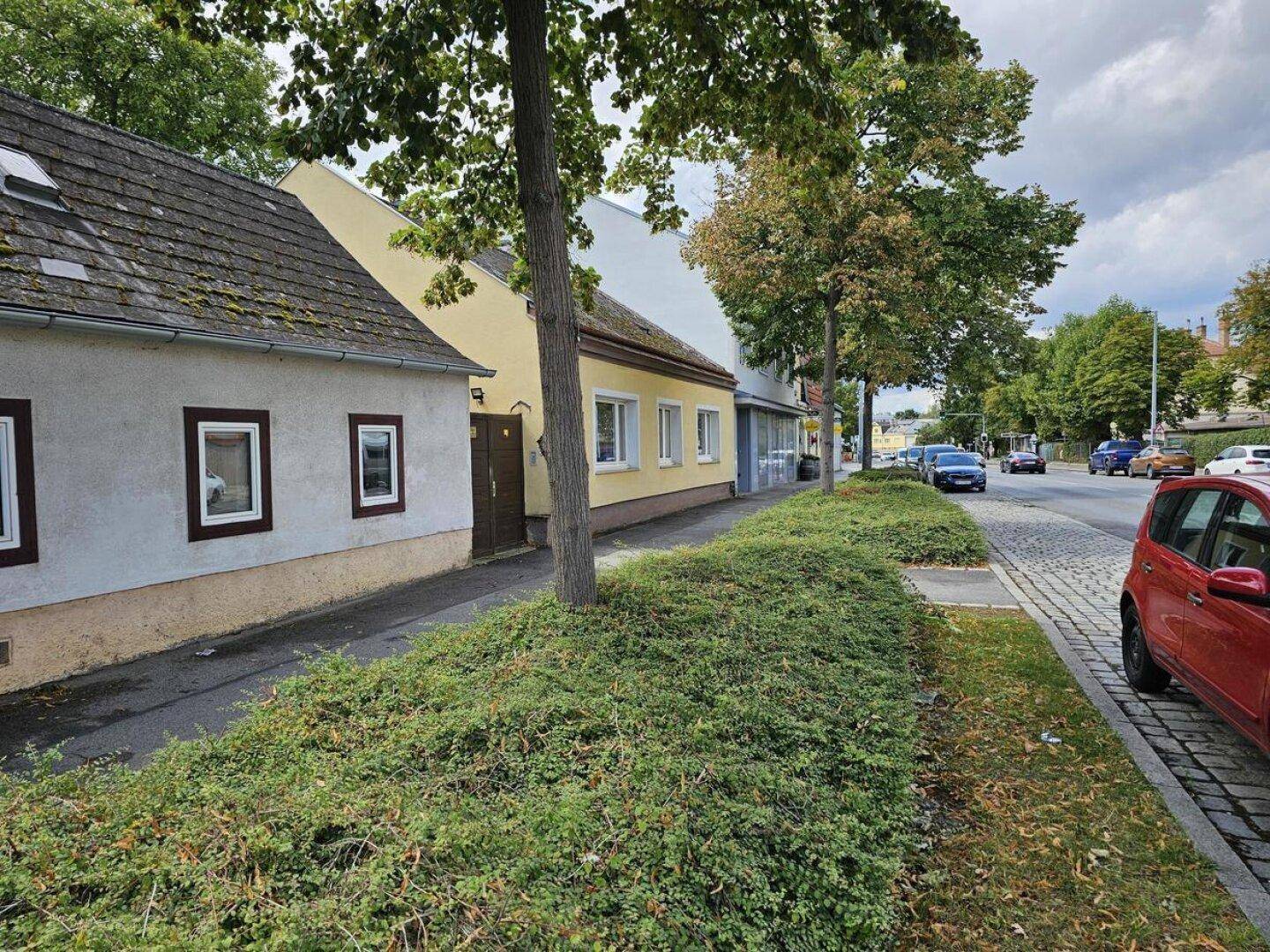 Straßenansicht mit dem Haus, Bäumen und parkenden Autos entlang der Fahrbahn an einem sonnigen Tag.
