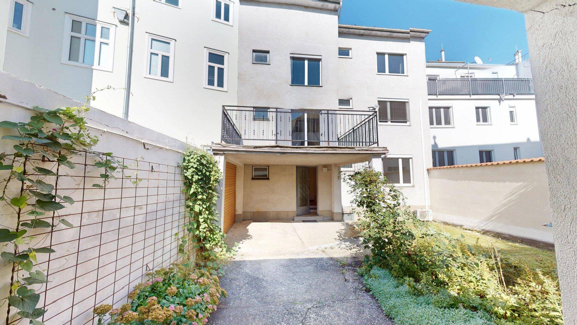 Rückseite des Gebäudes mit einem kleinen Balkon, Zufahrt und begrünten Bereichen.