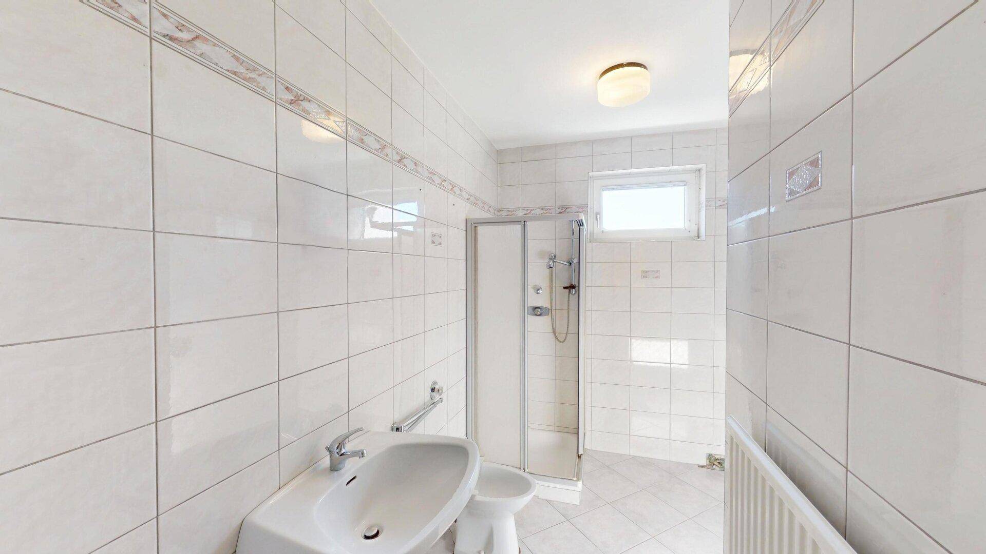 Gefliestes Badezimmer mit Dusche, Waschbecken und Fenster, in gutem Zustand.
