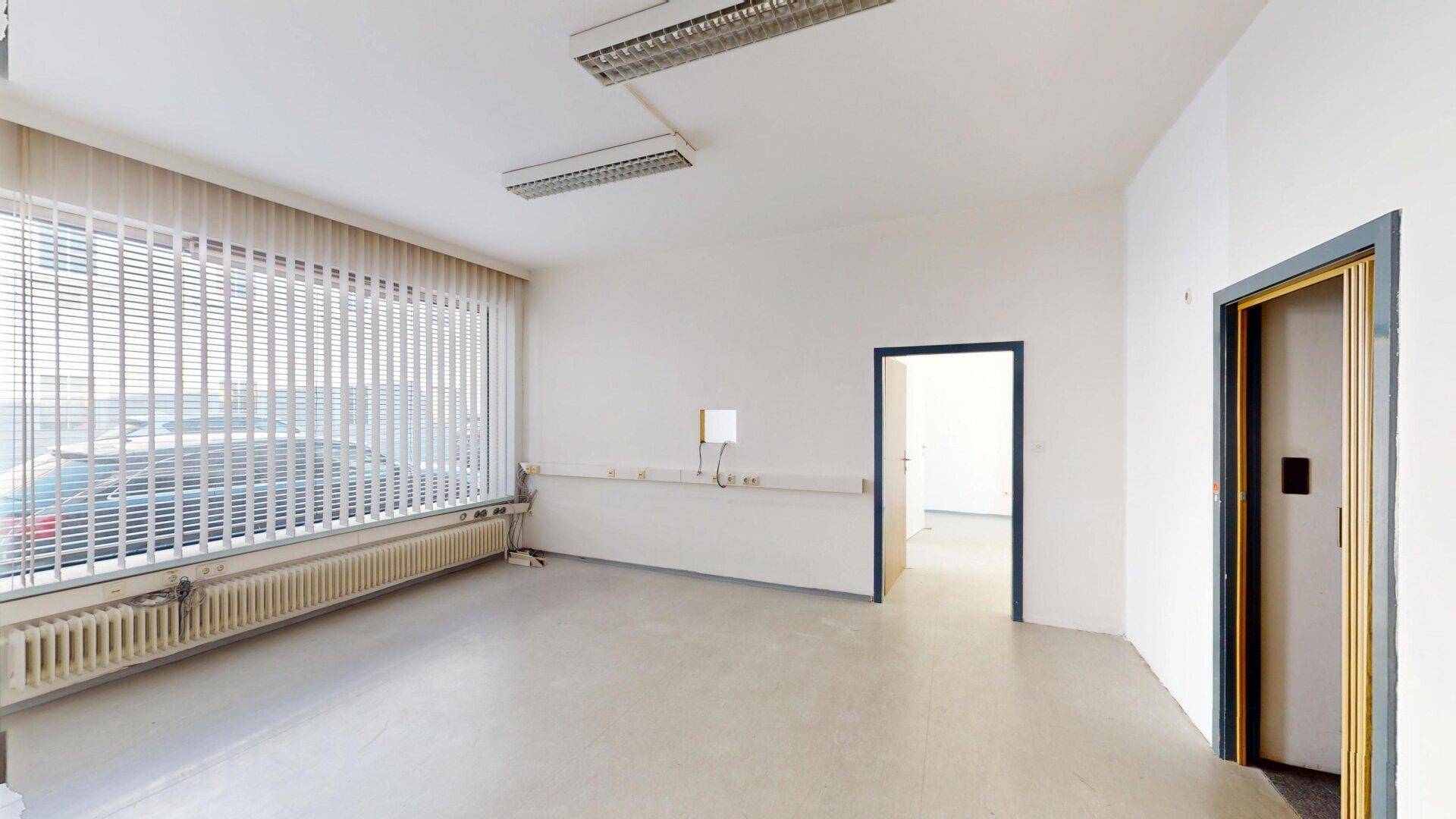 Heller Büroraum mit großem Fenster zur Straße und Jalousien für Privatsphäre.