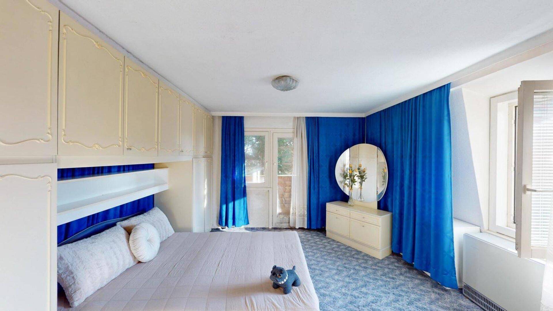 Schlafzimmer mit blauen Vorhängen, Einbauschränken und einem Schminktisch mit Spiegel.