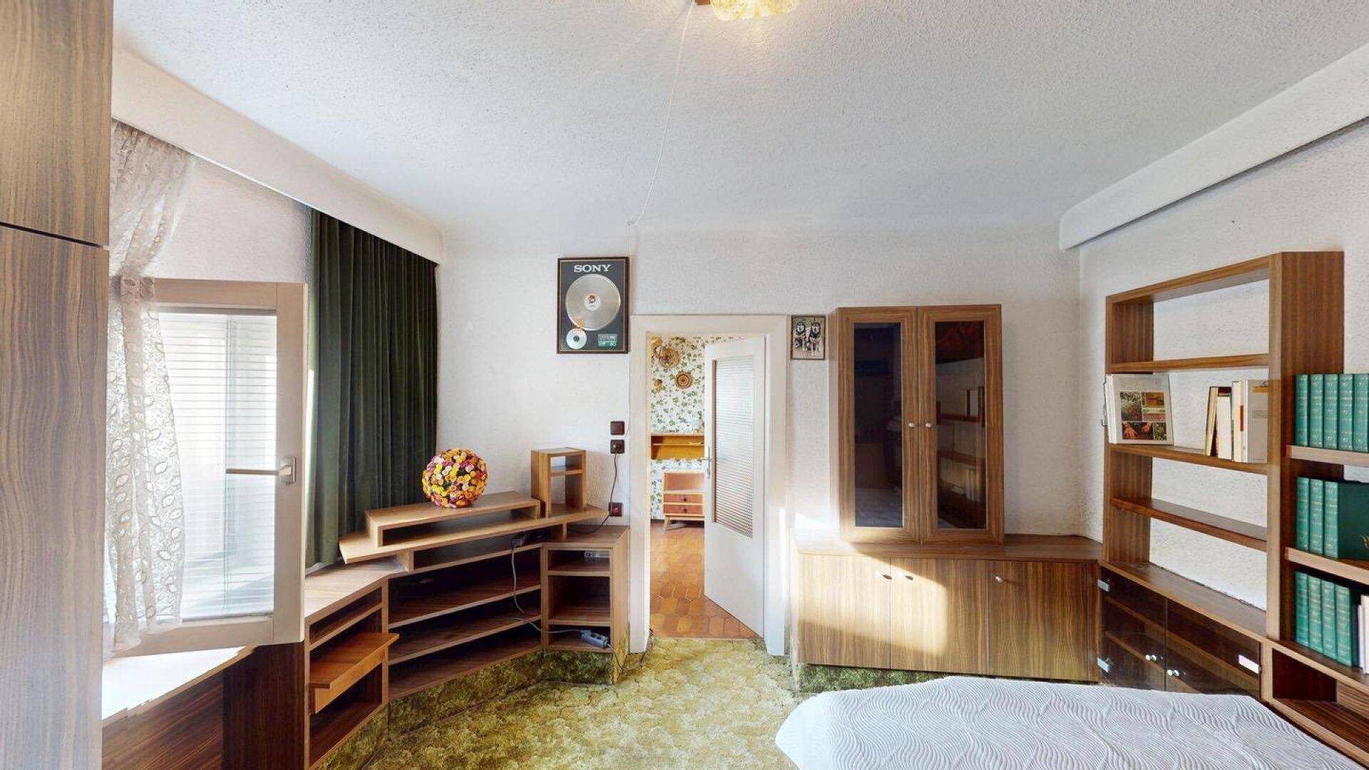 Schlafzimmer mit grünem Teppichboden, Holzmöbeln und einem Fenster mit Blick nach draußen.
