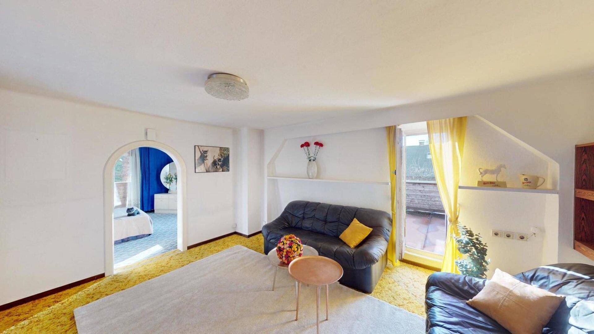 Wohnzimmer mit gelbem Teppichboden und einem Durchgang zu einem angrenzenden Schlafzimmer.