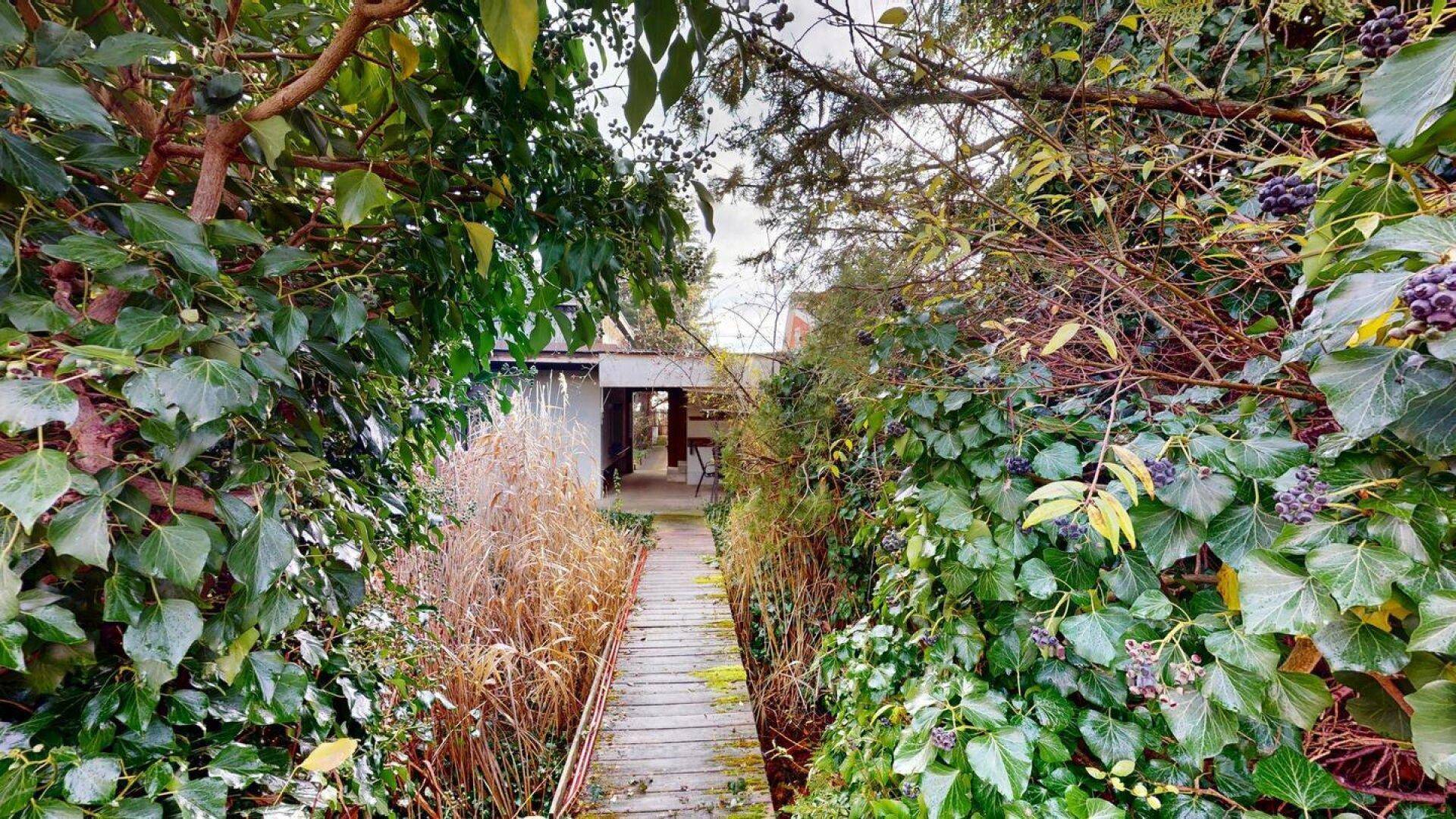 Holzsteg führt durch einen üppig bewachsenen Gartenbereich zu einem Gebäude.