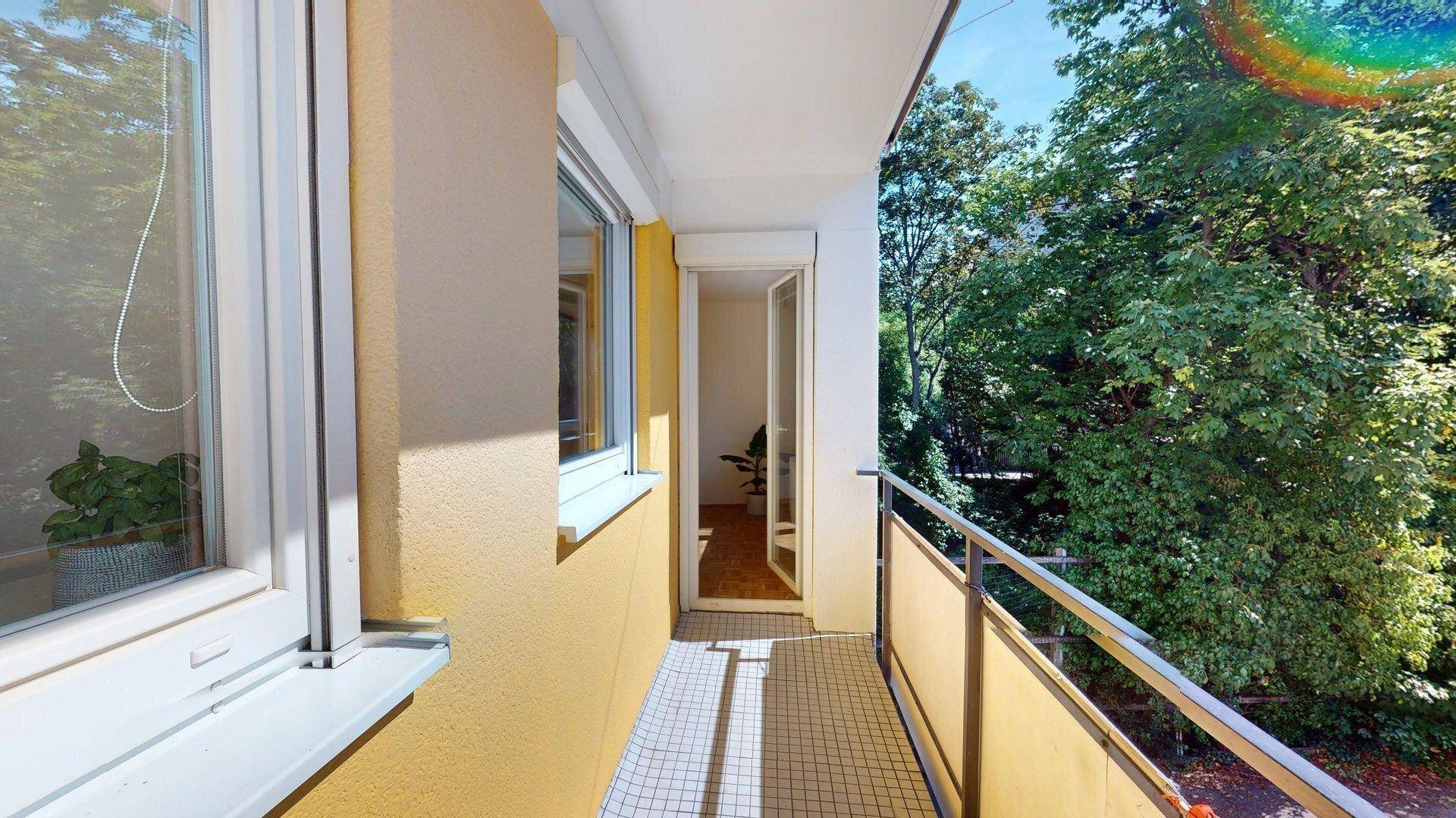 Heller Balkon mit Fliesenboden und Metallgeländer, umgeben von grünen Bäumen, bietet Zugang zum Innenbereich.