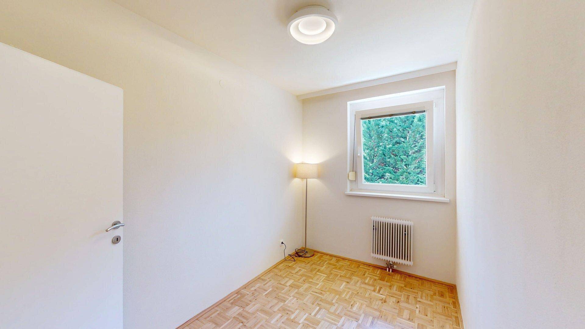 Helles Zimmer mit Parkettboden, einem Fenster mit Grünblick und einem Heizkörper.