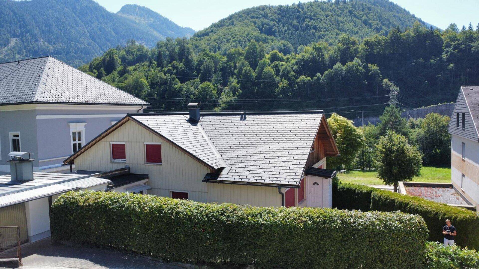 Seitenansicht des Hauses mit Satteldach, grüner Hecke und bewaldeten Bergen im Hintergrund.