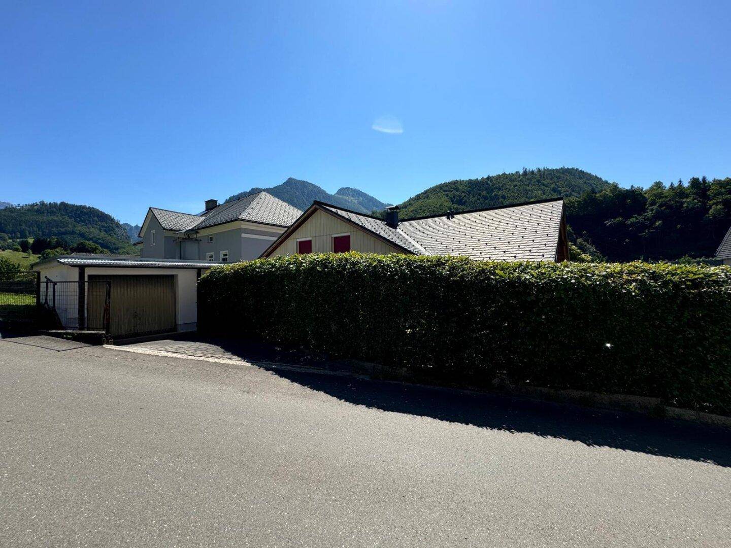 Blick von der Straße auf das Grundstück mit langer Hecke, Garage und Bergen im Hintergrund.