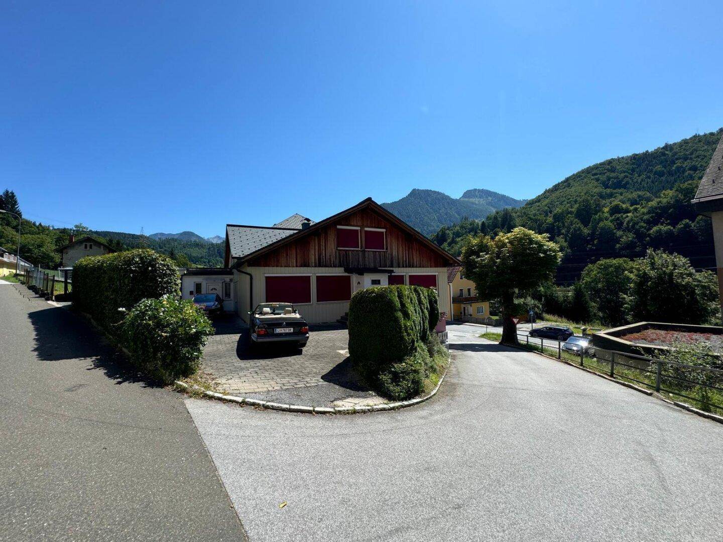 Vorderansicht des Hauses mit gepflasterter Einfahrt, Hecke und malerischer Berglandschaft.