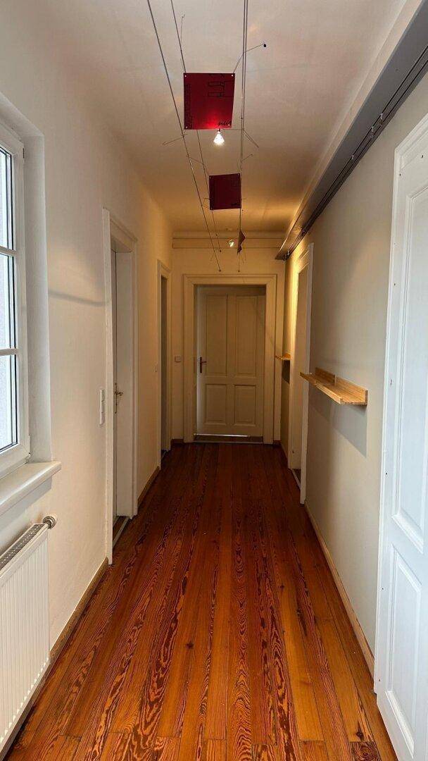 Langer Flur mit Holzdielenboden, roten Deckenleuchten und einem Fenster mit Heizkörper.