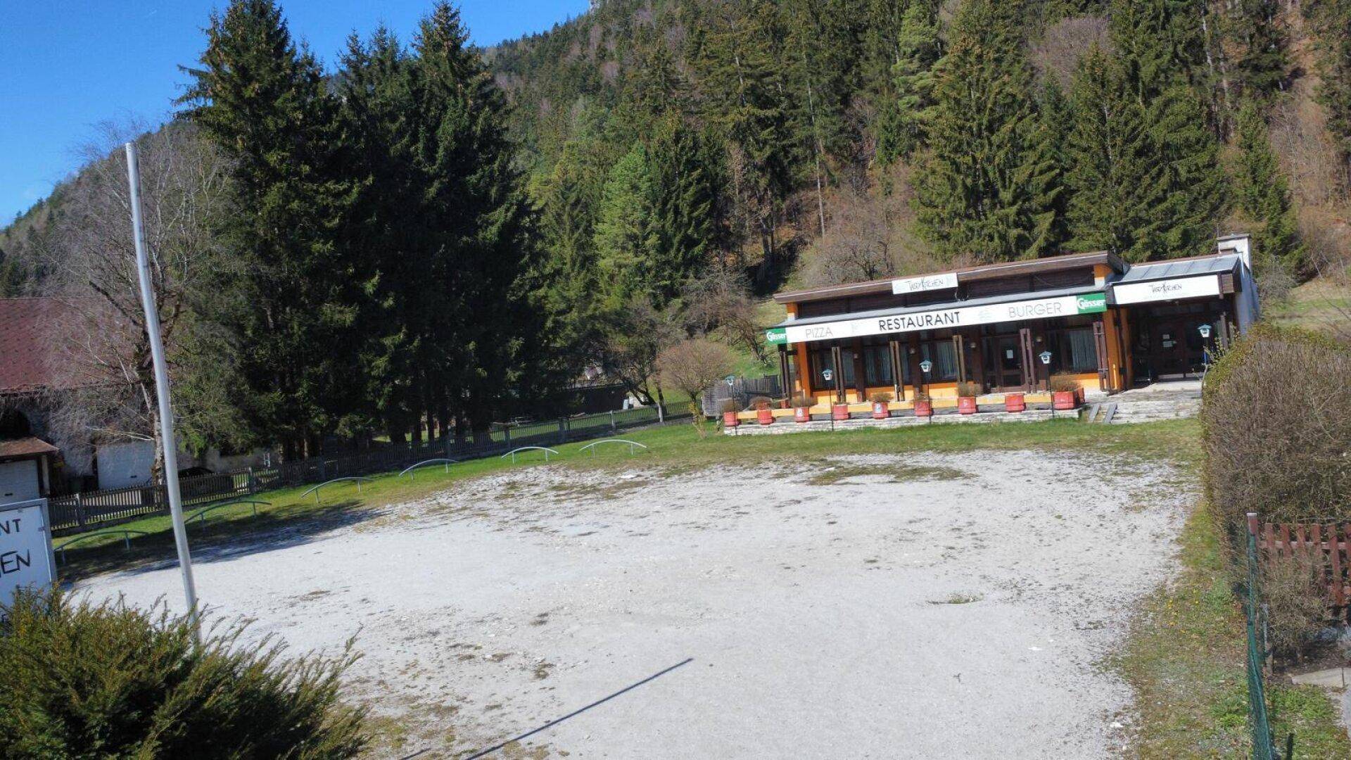 Restaurantgebäude mit großer Schotterfläche und dichtem Wald im Hintergrund unter blauem Himmel.