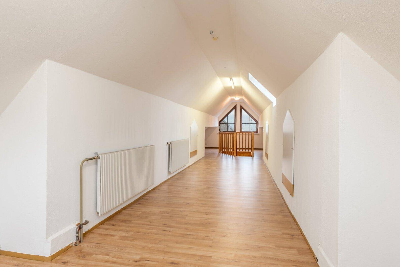 Langer Dachbodenraum mit Laminatboden, Heizkörpern und einem Giebelfenster am Ende.