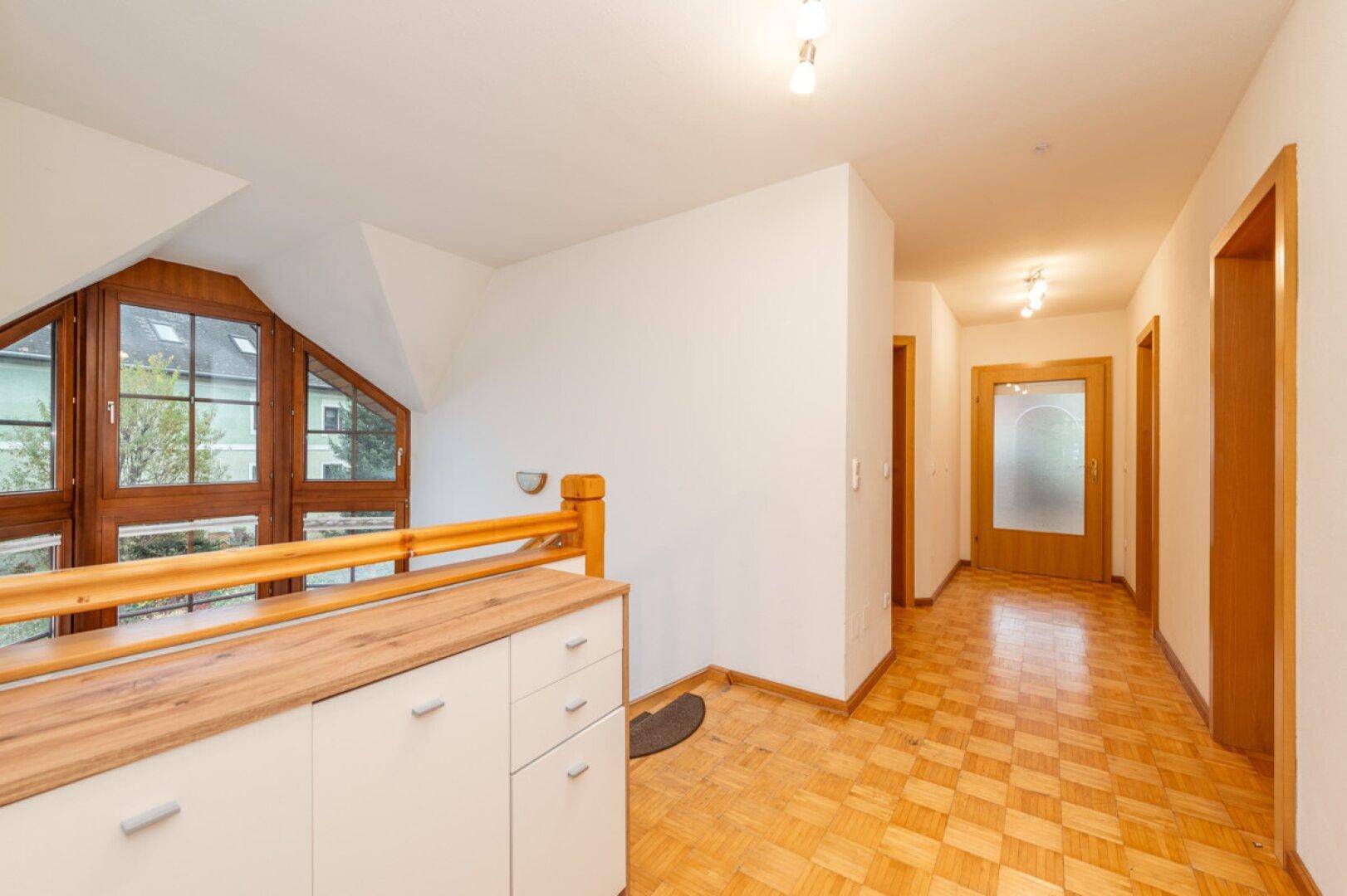 Heller Flur im Obergeschoss mit Parkettboden, niedrigem Schrank und großem Fenster.