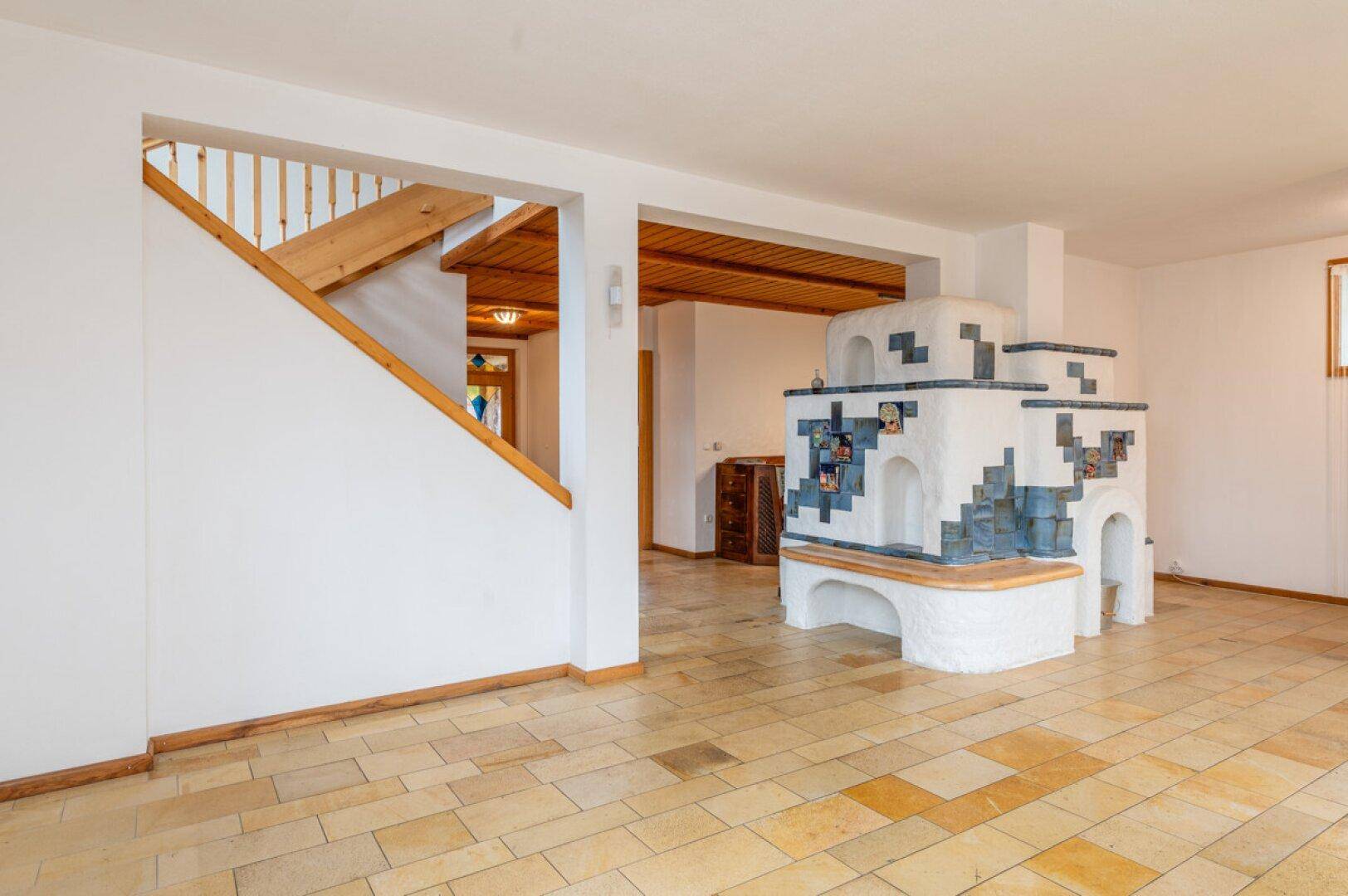 Geräumiger Wohnbereich mit traditionellem Kachelofen, Fliesenboden und Holztreppe zum Obergeschoss.