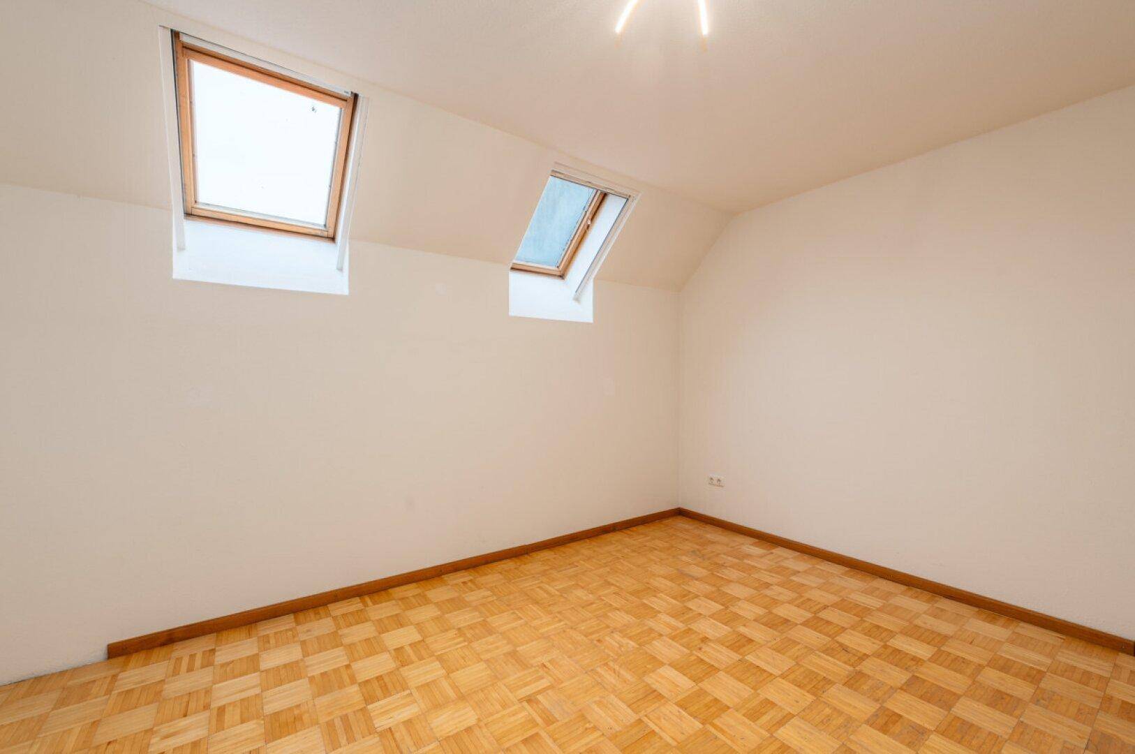 Geräumiges Zimmer mit Parkettboden, Dachschräge und zwei hellen Dachfenstern.