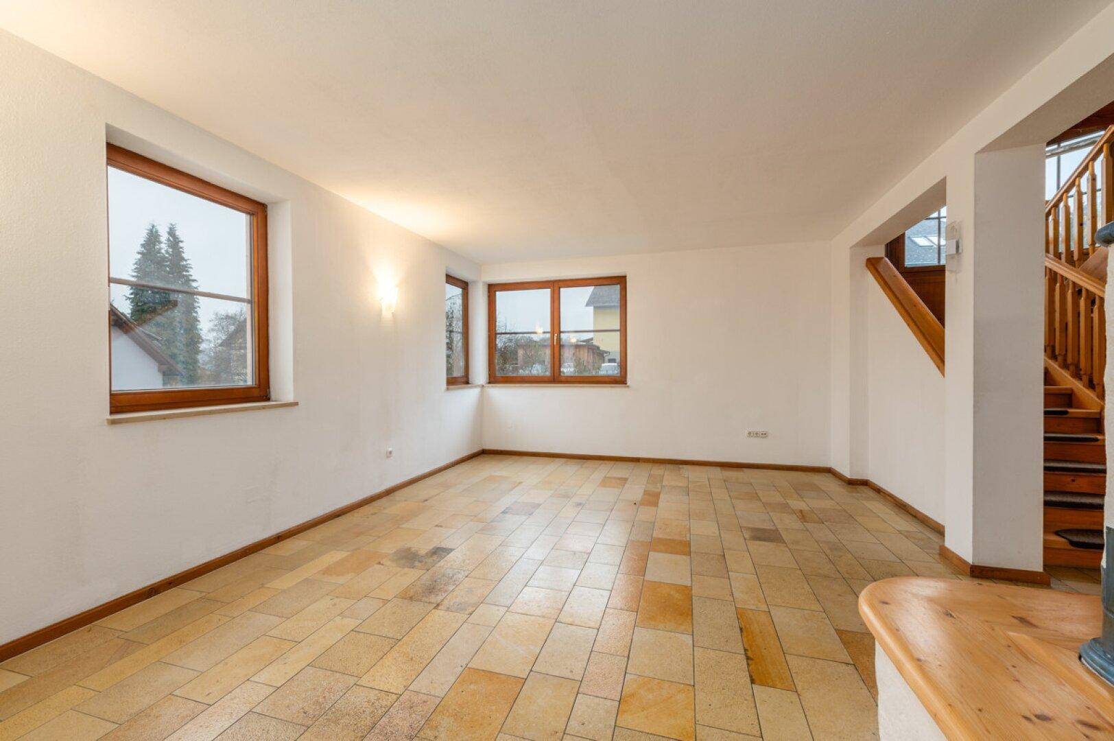 Heller, leerer Wohnraum mit Fliesenboden und großen Fenstern, angrenzend an eine Treppe.