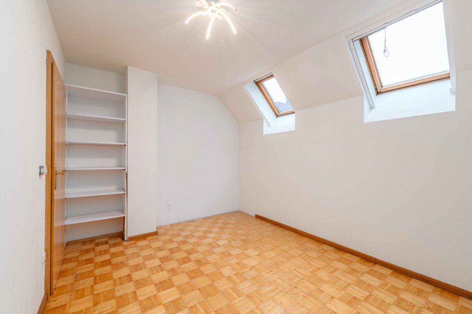 Helles Zimmer mit Parkettboden, Dachschräge, Regal und zwei Dachfenstern.