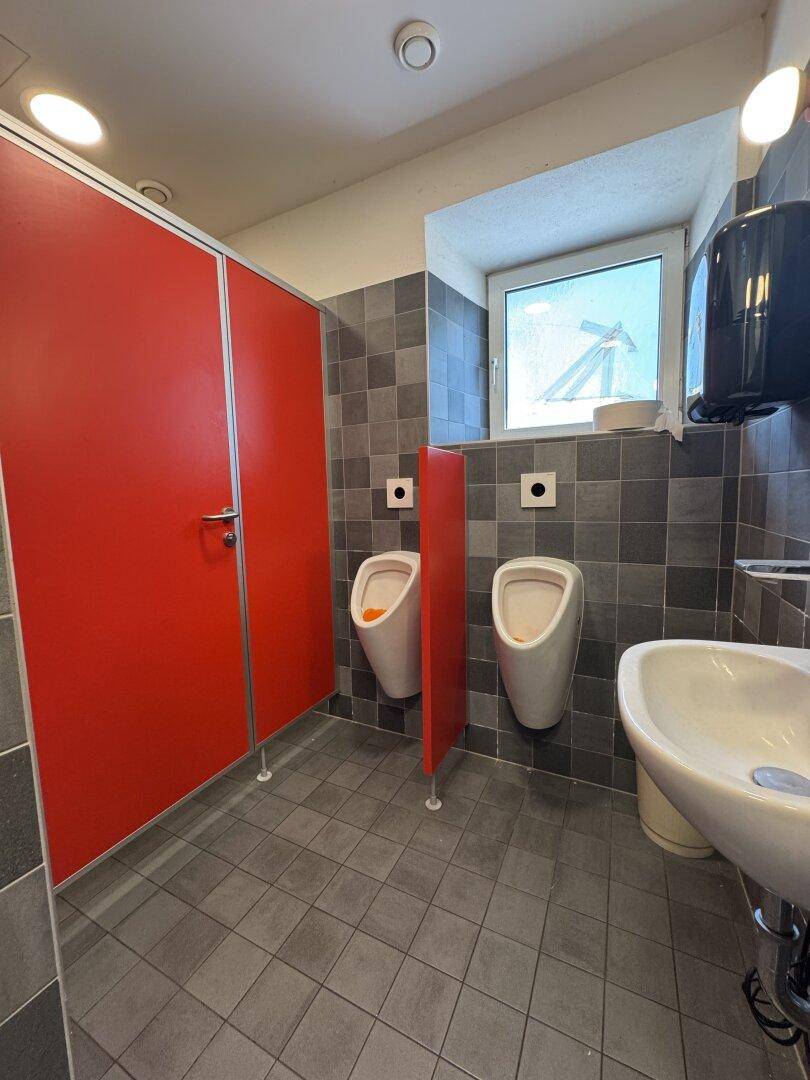 Herren-WC mit Urinalen, roten Kabinentüren und einem Fenster für Tageslicht.