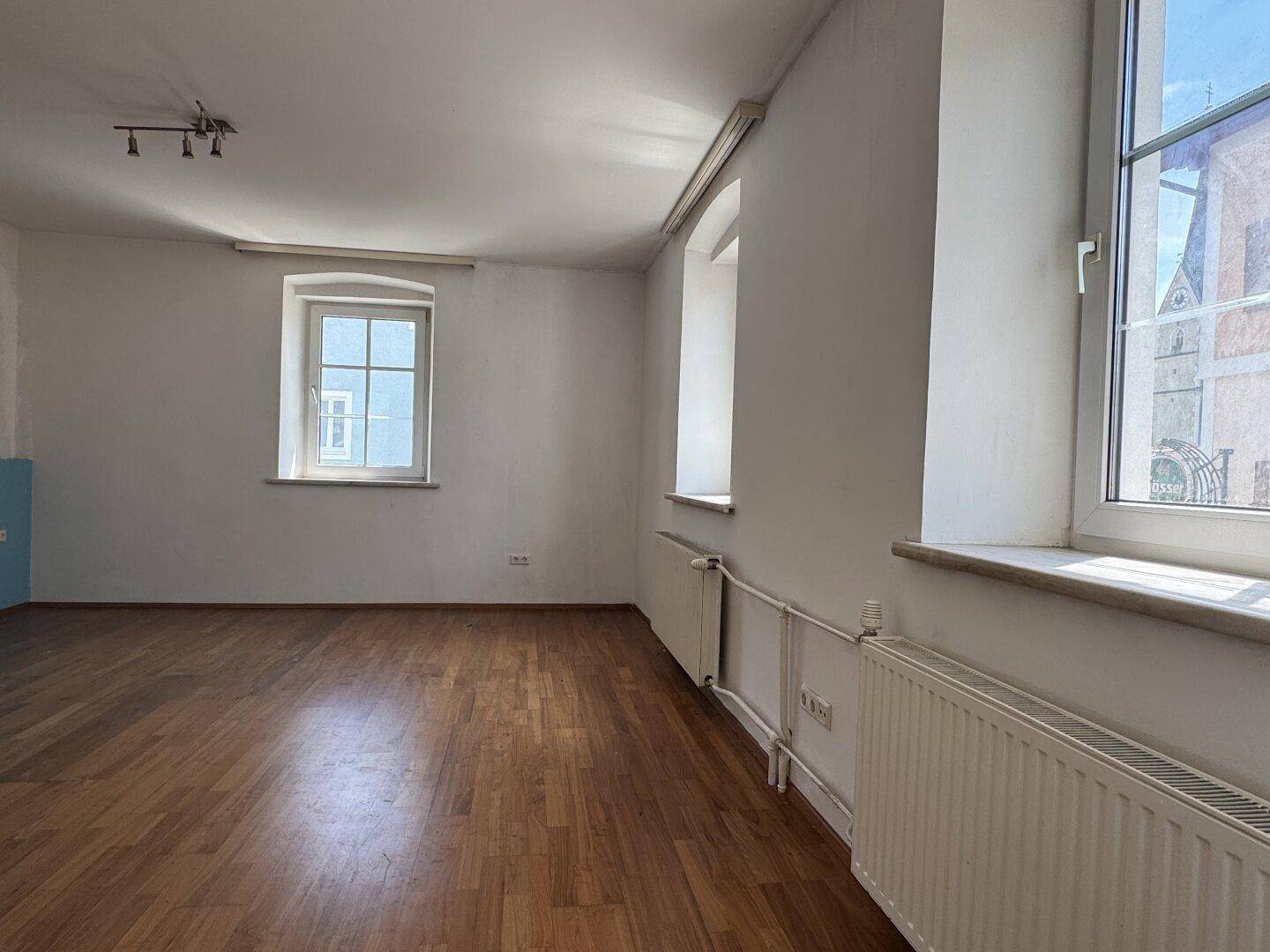 Leerer Raum mit Holzboden, weißen Wänden und großen Fenstern, die viel Licht hereinlassen.