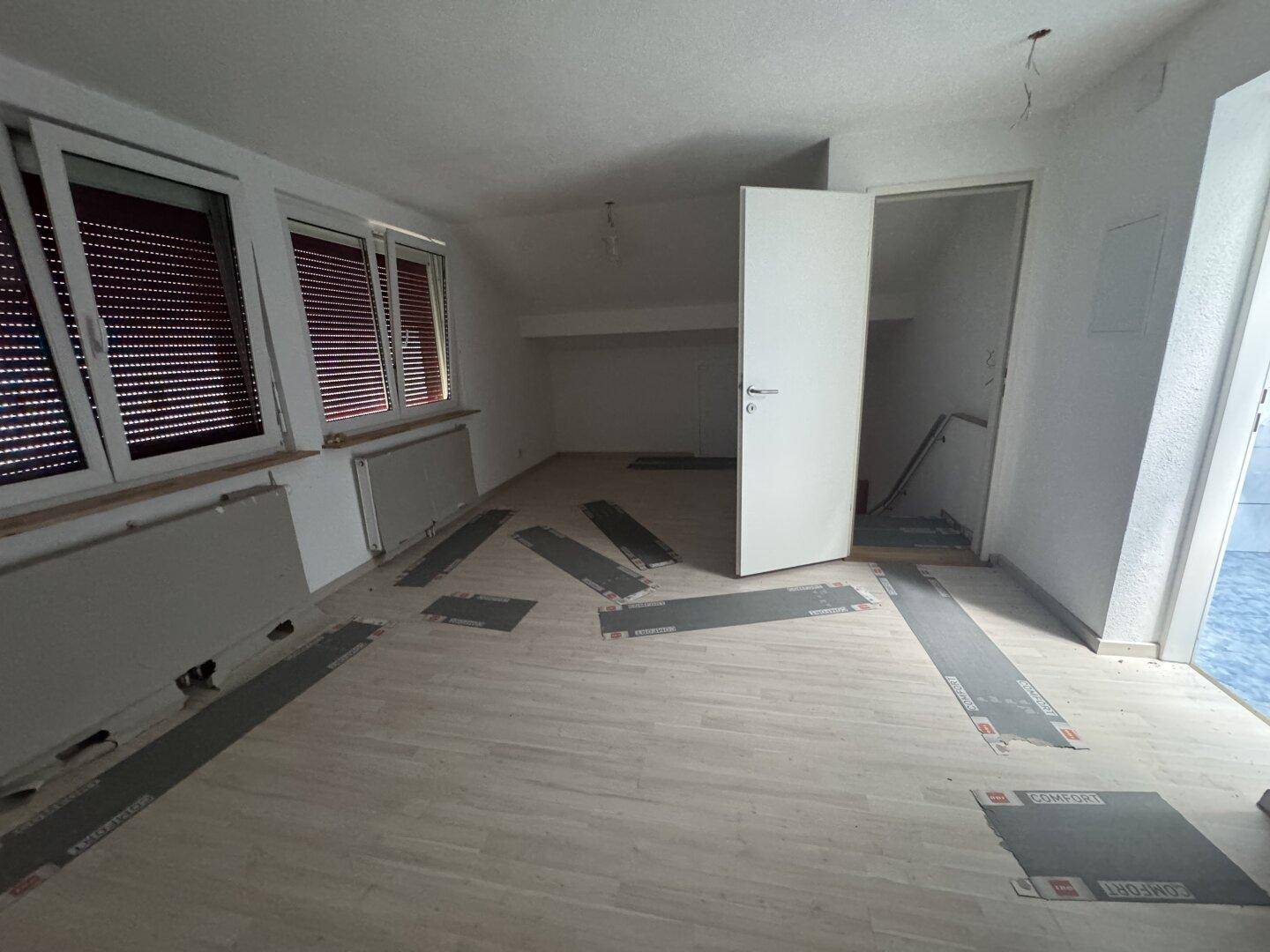 Leerer Raum im Dachgeschoss mit schrägen Wänden, mehreren Fenstern und teilweise verlegtem Bodenbelag.