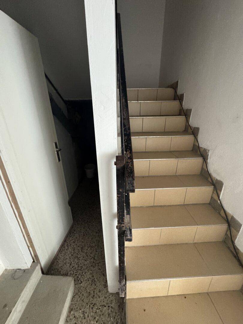 Treppenhaus mit beigefarbenen Fliesenstufen und dunklem Metallgeländer, führt zu einem Flur.