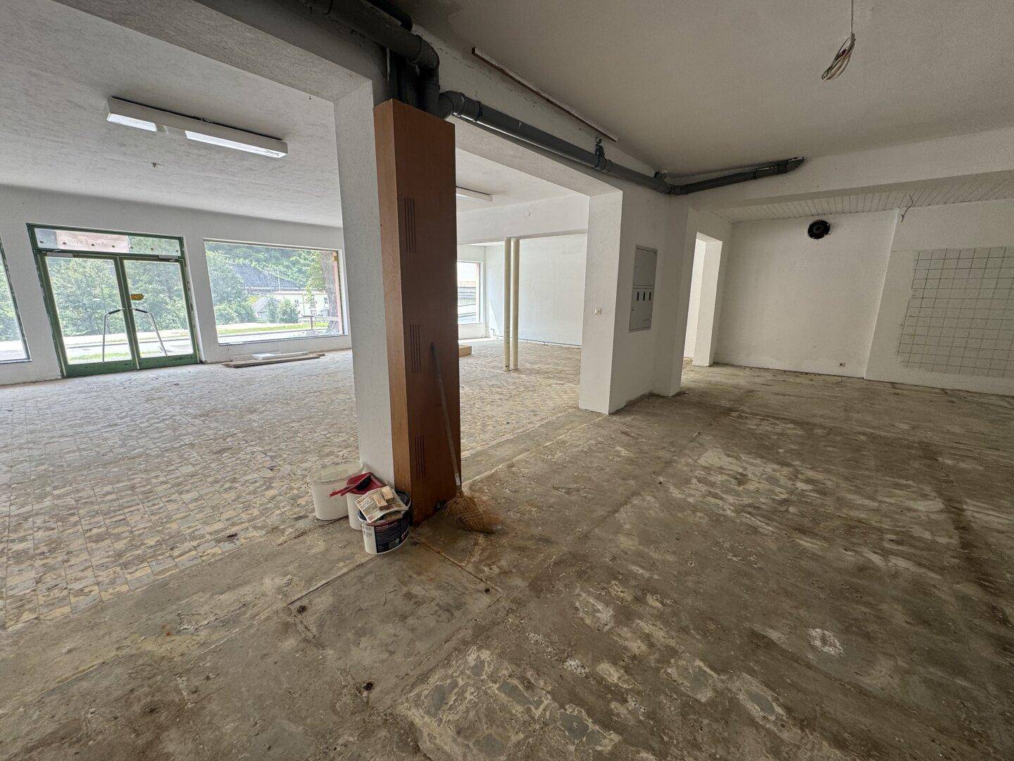 Großer, leerer Raum mit Betonboden, sichtbaren Rohren an der Decke und großen Schaufenstern.