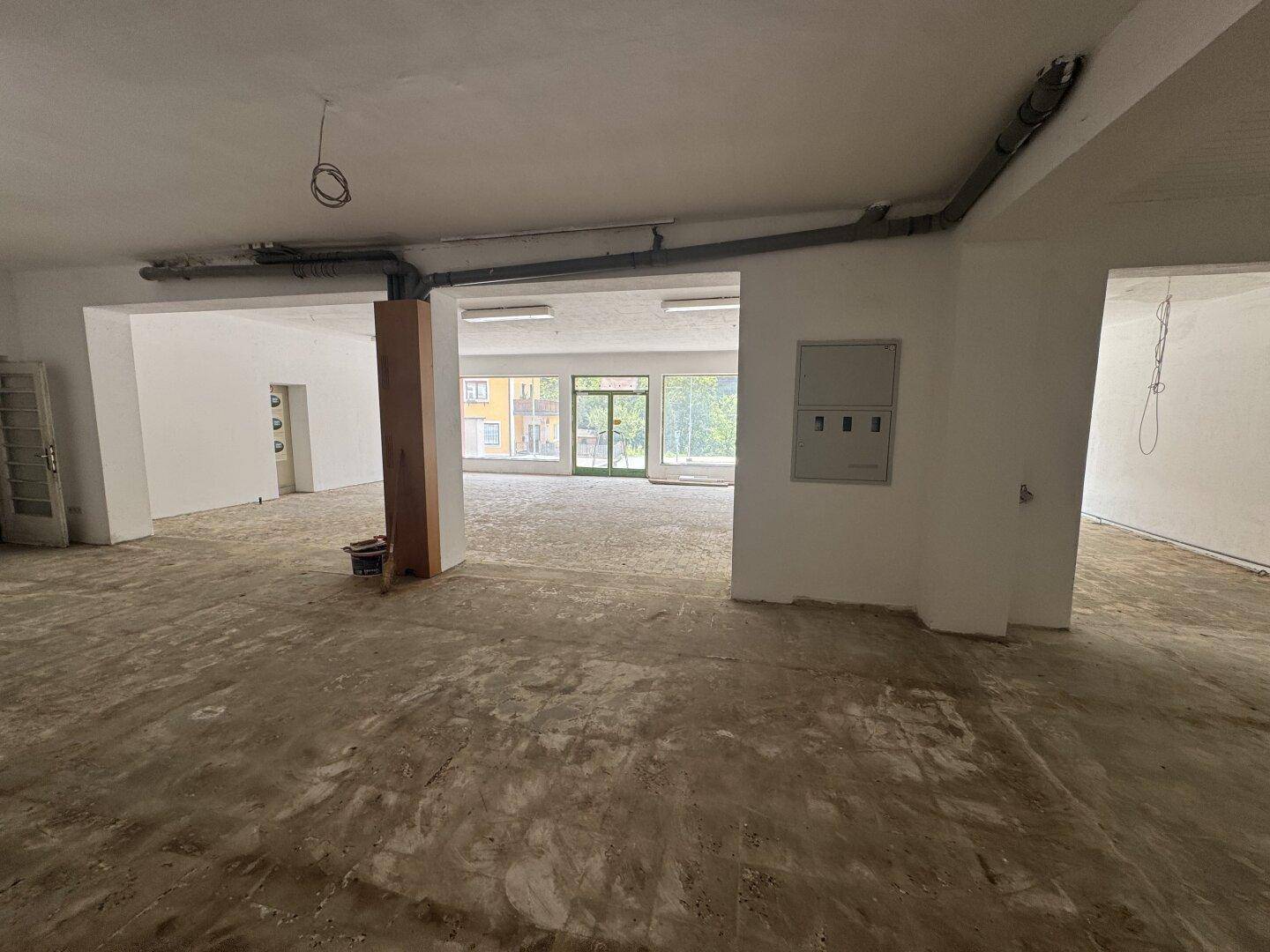 Weitläufiger, unmöblierter Raum mit Betonboden, hohen Decken und viel Tageslicht durch große Fenster.