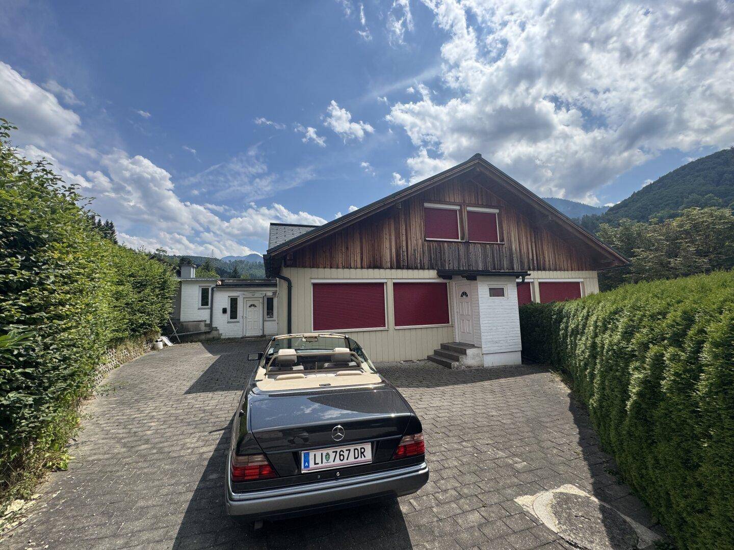 Haus mit Holzfassade und roten Fensterläden, gepflasterter Auffahrt und Bergblick im Hintergrund.