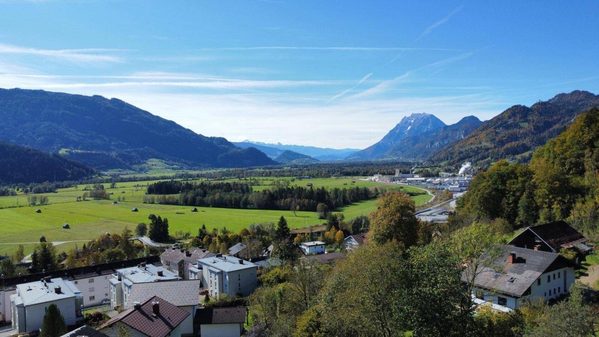 Weitläufiger Blick über eine ländliche Gegend mit Häusern, grünen Feldern und Bergen im Hintergrund.