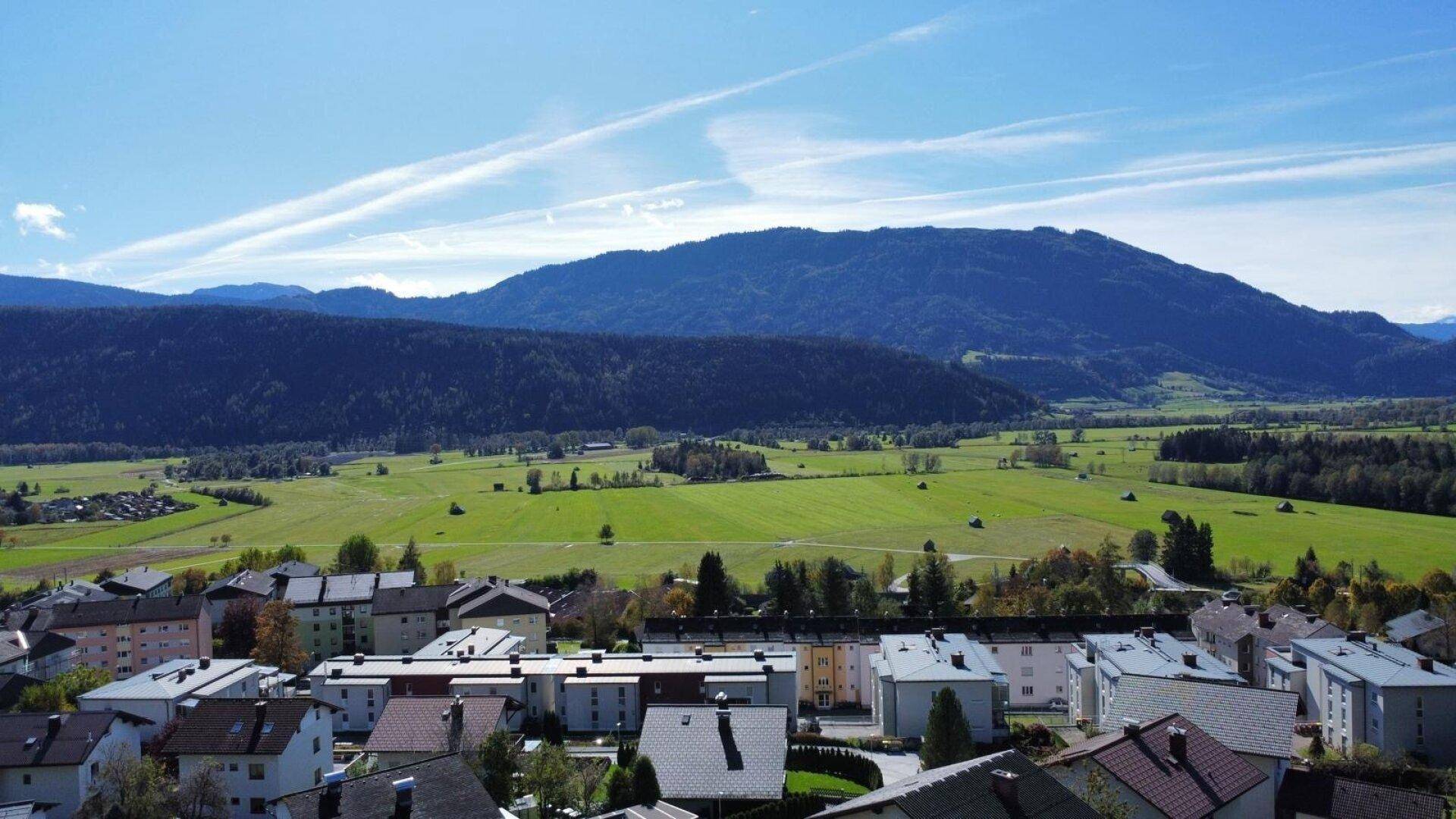 Panoramablick über eine Siedlung mit weiten grünen Wiesen und bewaldeten Bergen unter blauem Himmel.