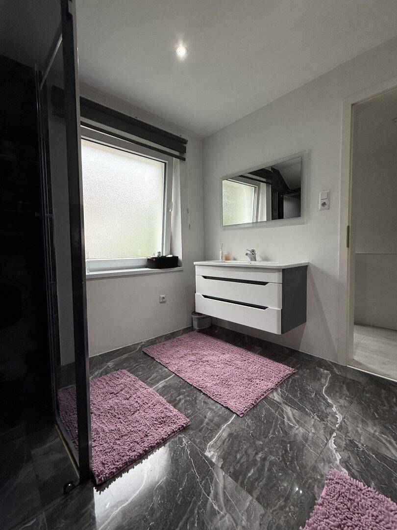 Helles Badezimmer mit modernem Waschtisch, Spiegel und Fenster, auf dunklen Marmorfliesen.