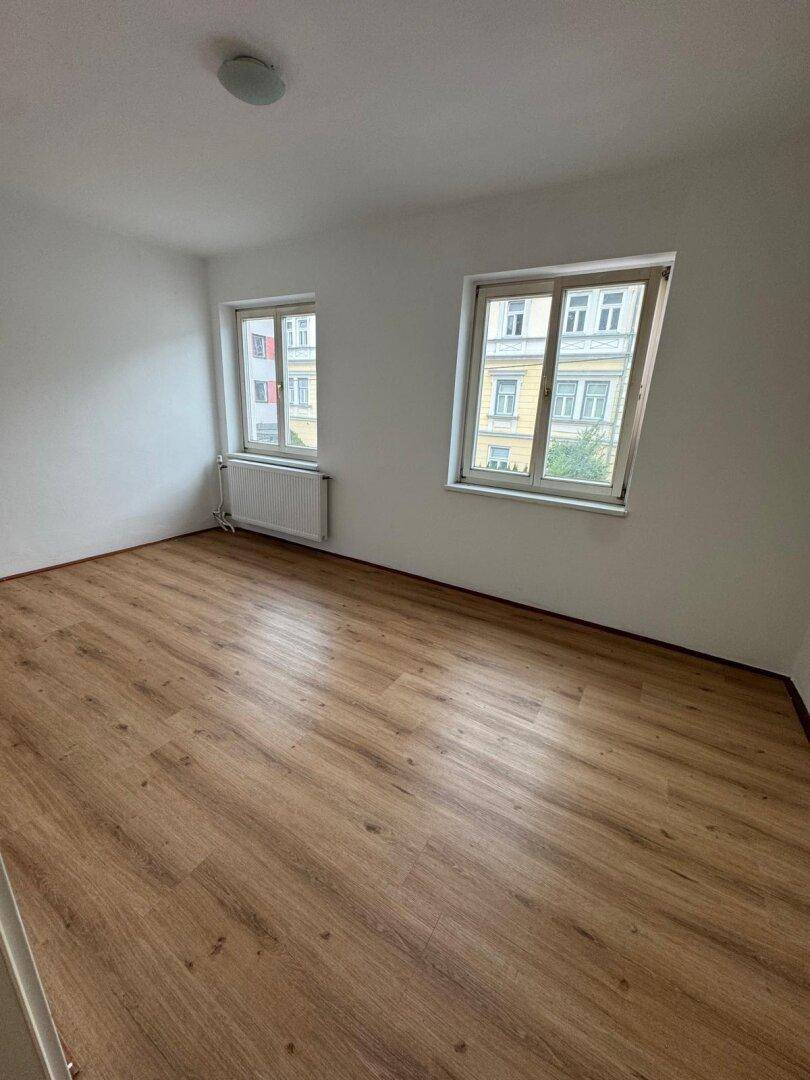 Leeres Zimmer mit Laminatboden und zwei Fenstern, die auf gegenüberliegende Gebäude blicken.