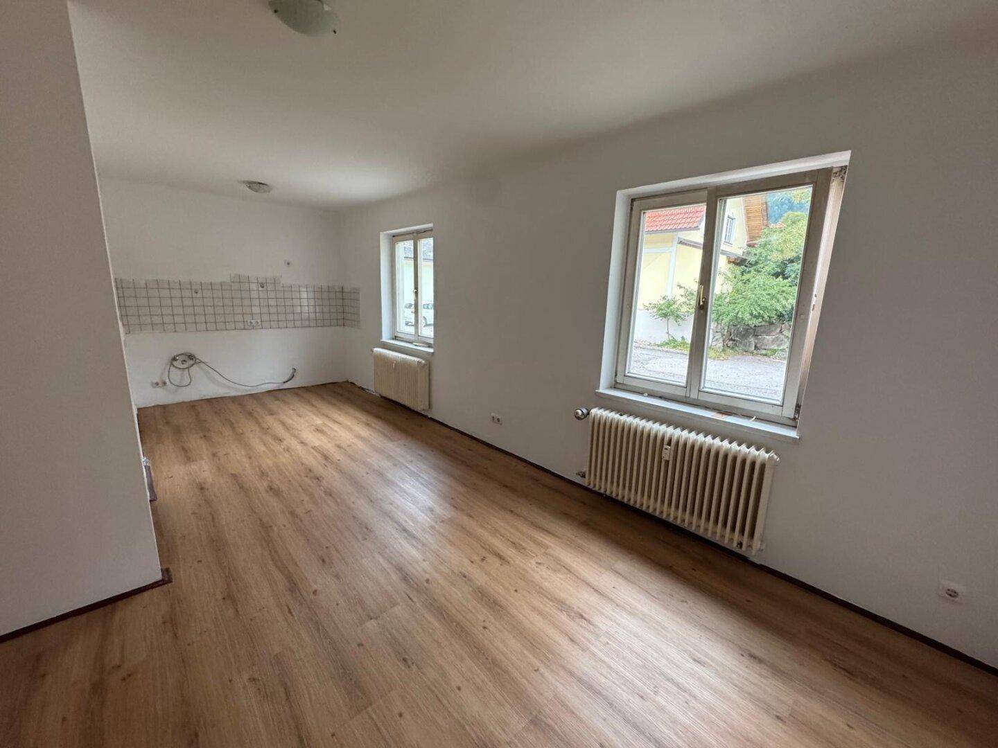 Leerer Raum mit Laminatboden und vorbereiteter Wand für eine Küche, mit zwei Fenstern.