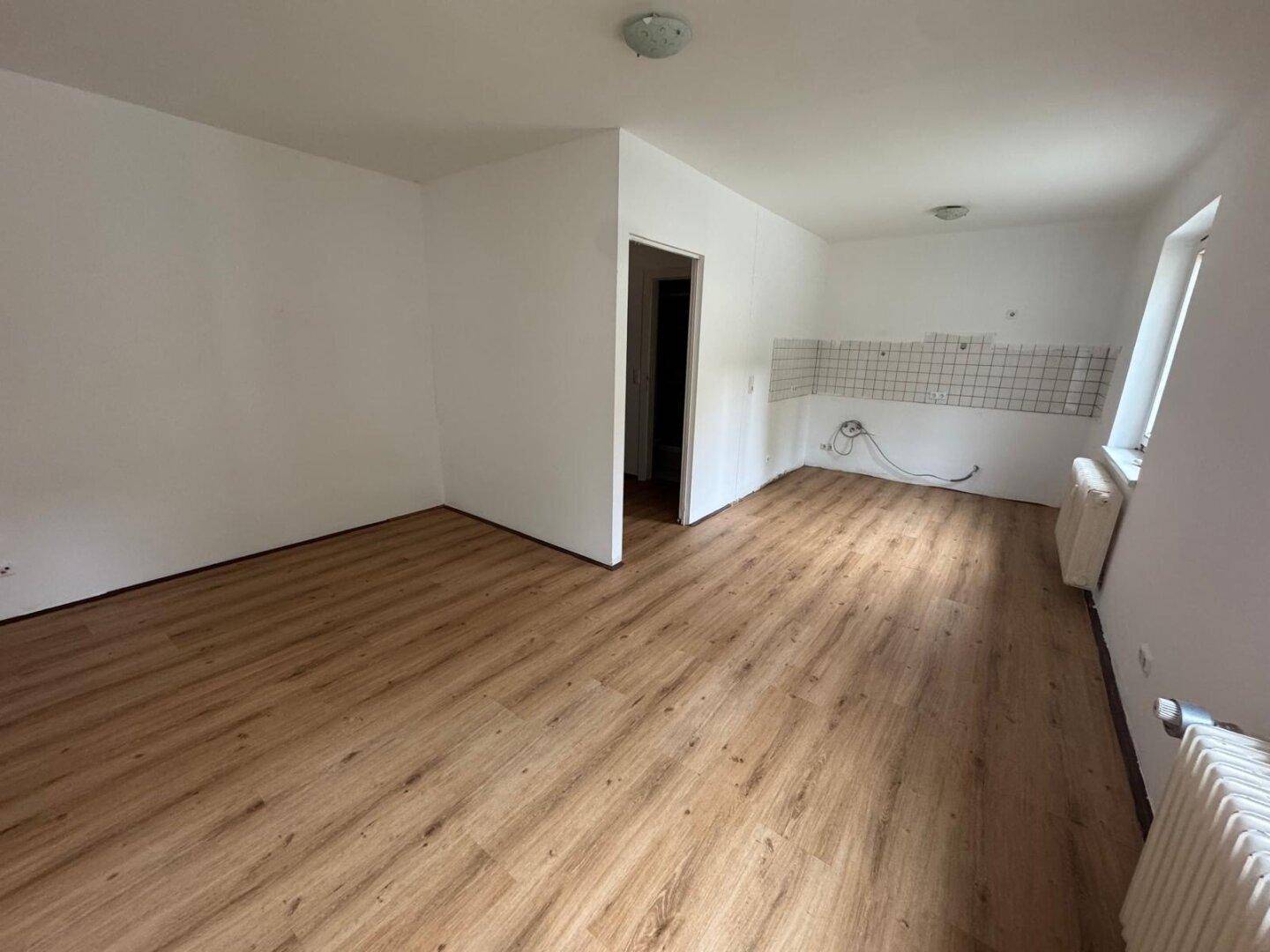 Leerer Raum mit Laminatboden und vorbereiteter Wand für eine Küche, mit Zugang zu einem weiteren Raum.