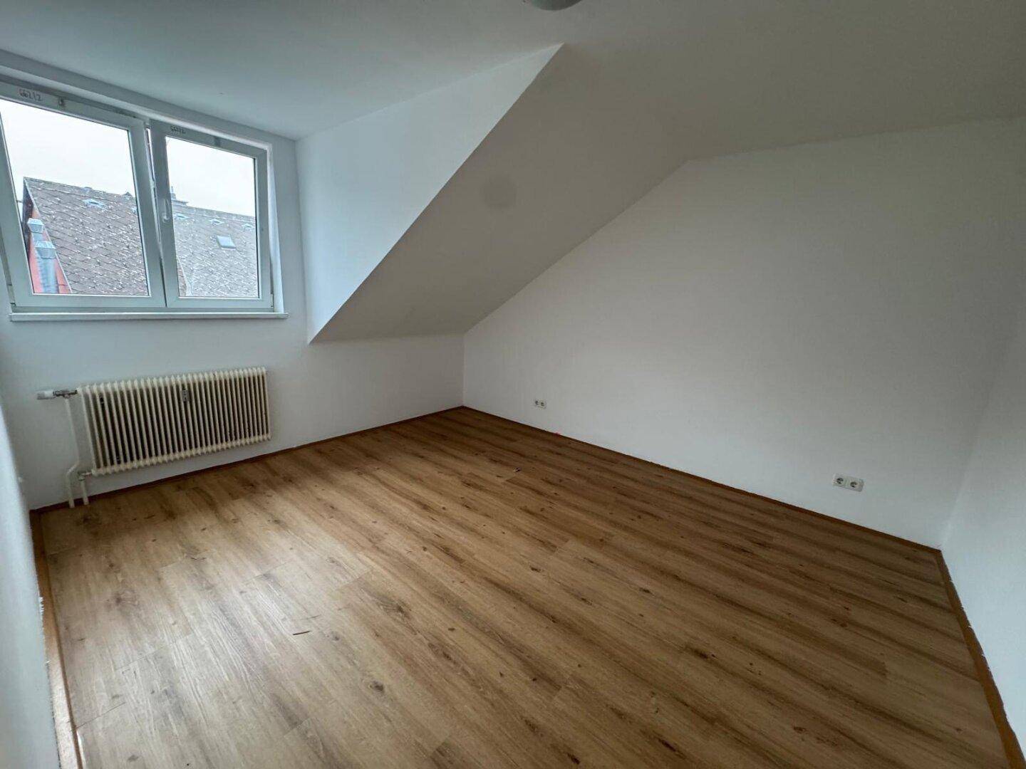 Helles Zimmer im Dachgeschoss mit Laminatboden, schräger Decke und einem Fenster.