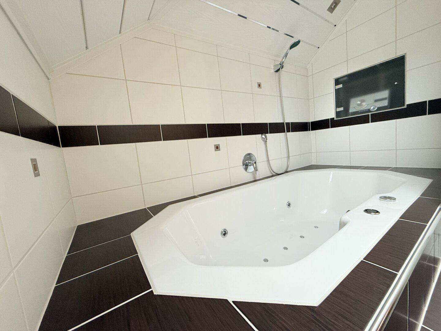 Geräumiges Badezimmer mit Whirlpool-Badewanne und dunklen Fliesen unter einer Dachschräge.