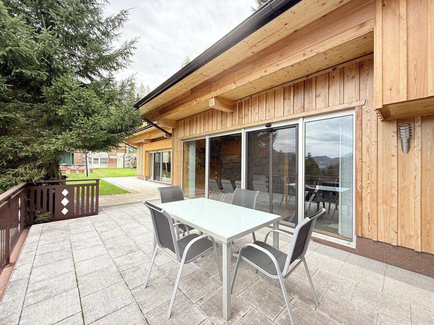 Möblierte Terrasse mit Steinplatten und Holzwänden, umgeben von Bäumen und Grünflächen.